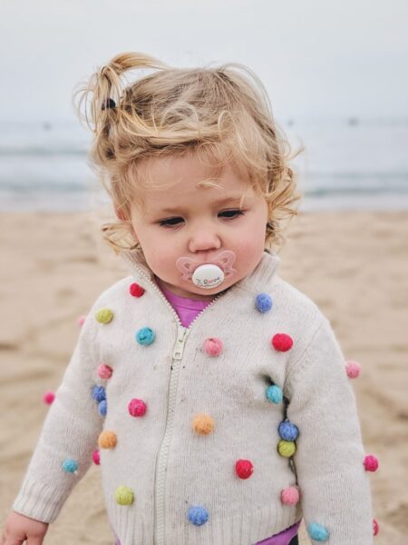 Ein kleines Kind mit Schnuller steht am Strand und trägt eine Jacke, auf der Bommel befestigt sind