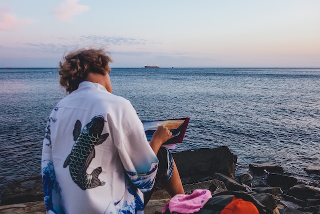 Eine Person sitzt mit dem Rücken zur Kamera und zeichnet das Meer. Sie trägt ein Hemd mit großem Fisch-Print.