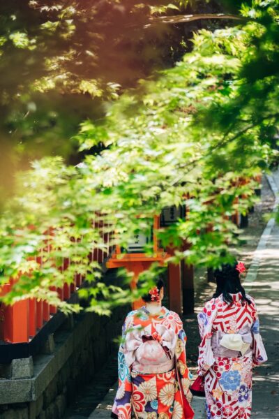 Zwei Frauen in traditioneller japanischer Kleidung laufen einen Weg entlanfg, im Vordergrund grüne Äste