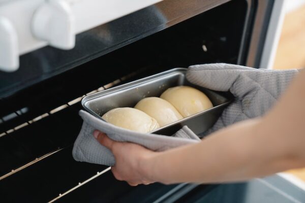 Hände mit Topflappen und Ofenhandschuh, die eine Auflaufform mit drei Teigkugeln in den Ofen stellen 