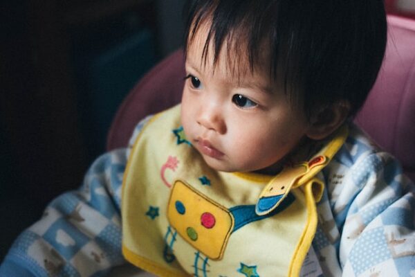 Ein Baby sitzt in einem Hochstuhl und trägt einen blauen Pullover und ein gelbes Lätzchen 