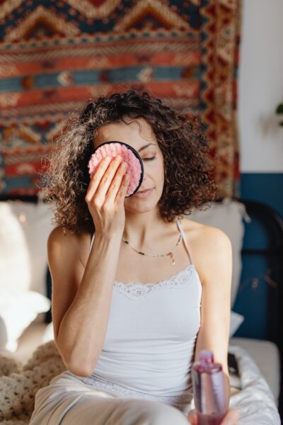 Eine Frau sitzt auf einem Bett und reinigt ihr Gesicht mit einem Abschminkpad aus rosa Stoff