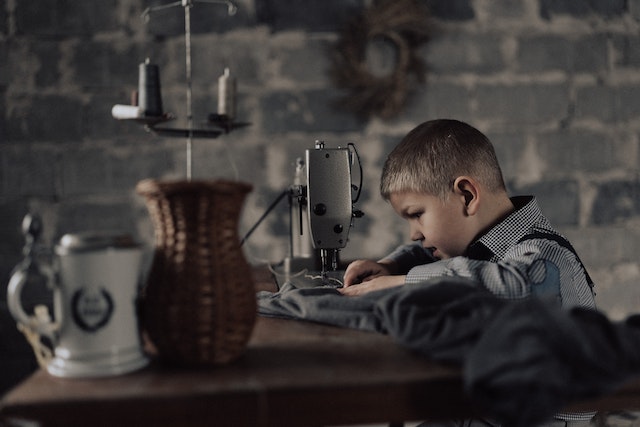 Ein Junge sitzt an einem Holztisch und näht an einer Nähmaschine, im Vordergrund steht ein Krug