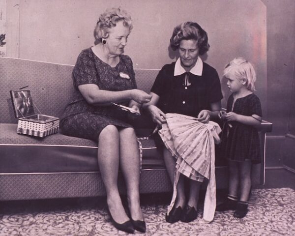 Zwei ältere Damen sitzen auf einem Sofa und nähen ein Kleid. Daneben steht ein Kind, dem eine der Frauen gerade eine Nadel zeigt 
